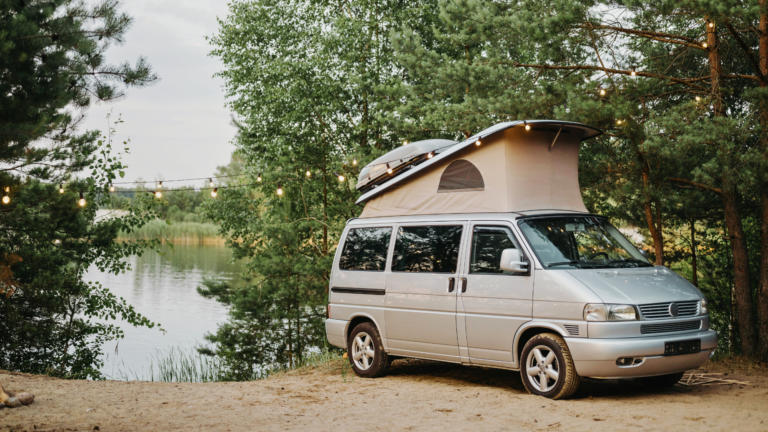 Van aménagé gris avec toit relevable, garé près d’un lac entouré de pins, sous une guirlande lumineuse suspendue entre les arbres.
