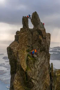A group of people climbing on the Svolvær Goat, 150-metre (490 ft), a jagged pinnacle high above the landscape of the Lofoten.