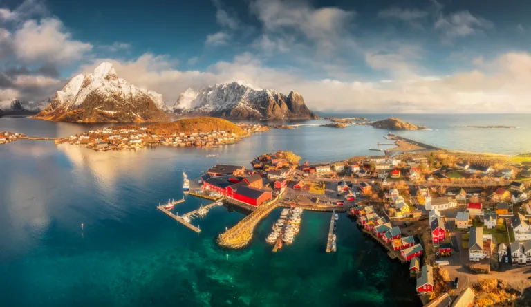 Panoramic aerial view of Reine fishing village with red rorbu houses at sunrise. Winter in Lofoten islands, Norway. Top drone view of snowy mountains, sea, reflection in water, blue sky with clouds